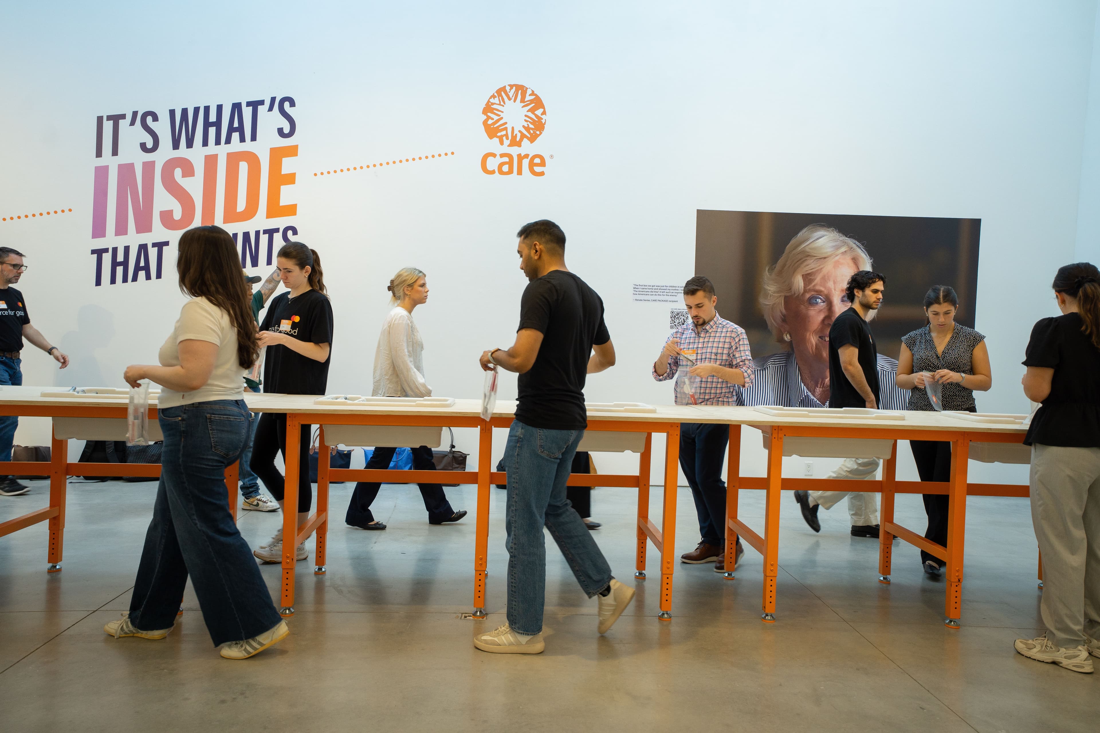 Visitors and volunteers assembling CARE PACKAGE kits at long tables beneath the "It's What's Inside That Counts" wall graphic and a large portrait photo