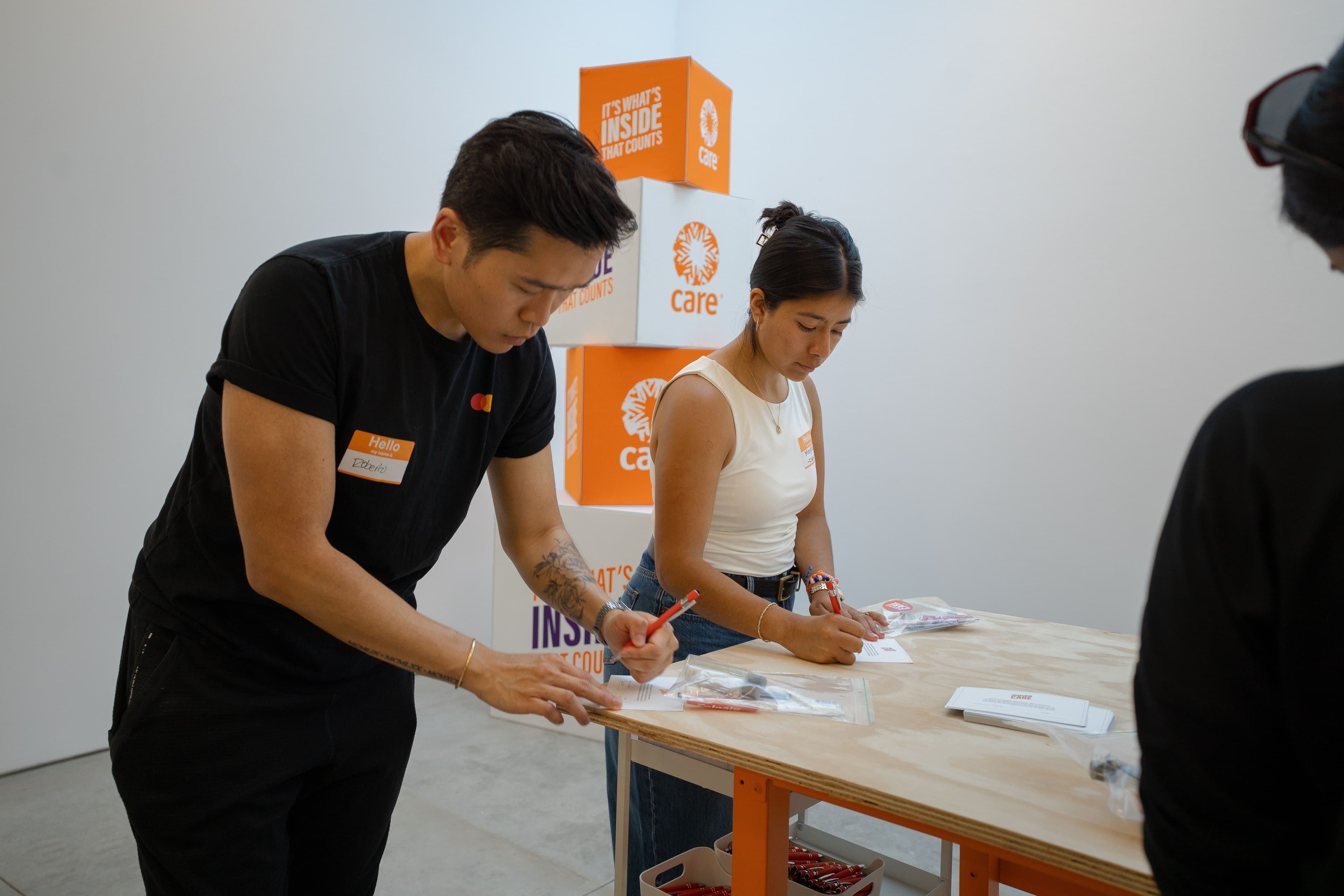 Two volunteers filling out CARE PACKAGE assembly cards at a wooden table, with stacked orange "It's What's Inside That Counts" boxes and the CARE logo behind them