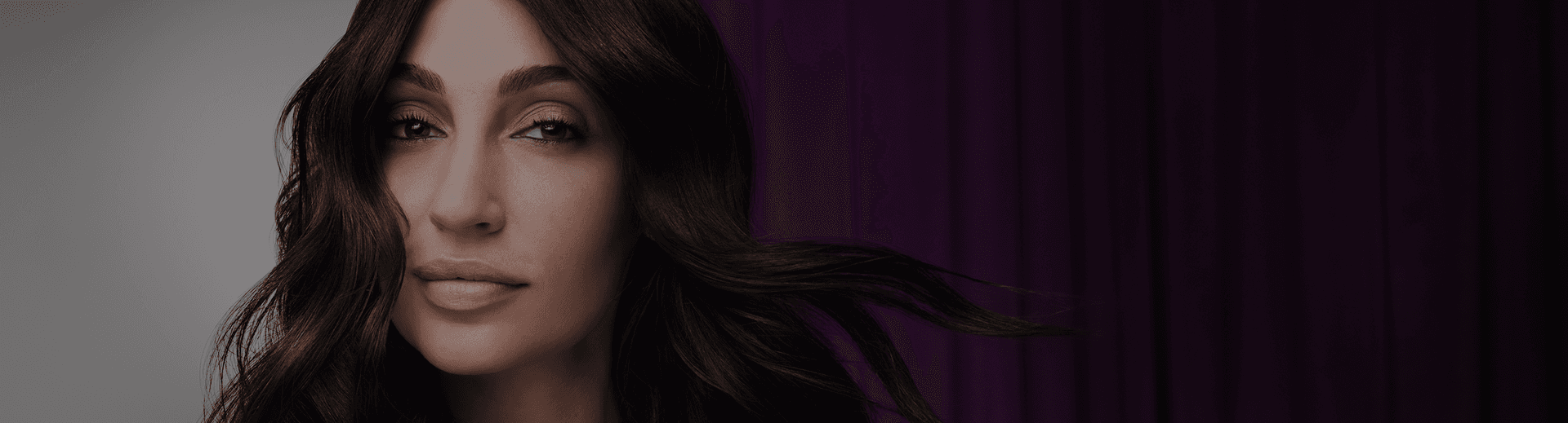 Woman with long, glossy dark brunette waves, hair color result of Wella Koleston, against a deep purple curtain backdrop.