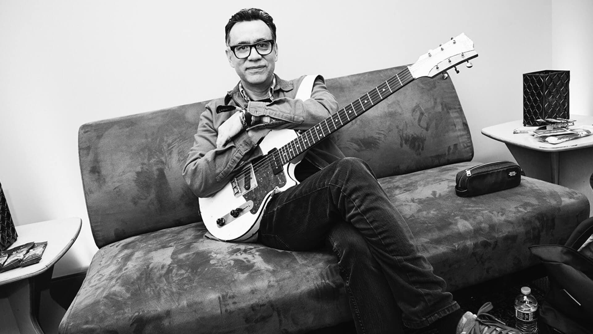 Fred Armisen sitting on a couch holding a guitar
